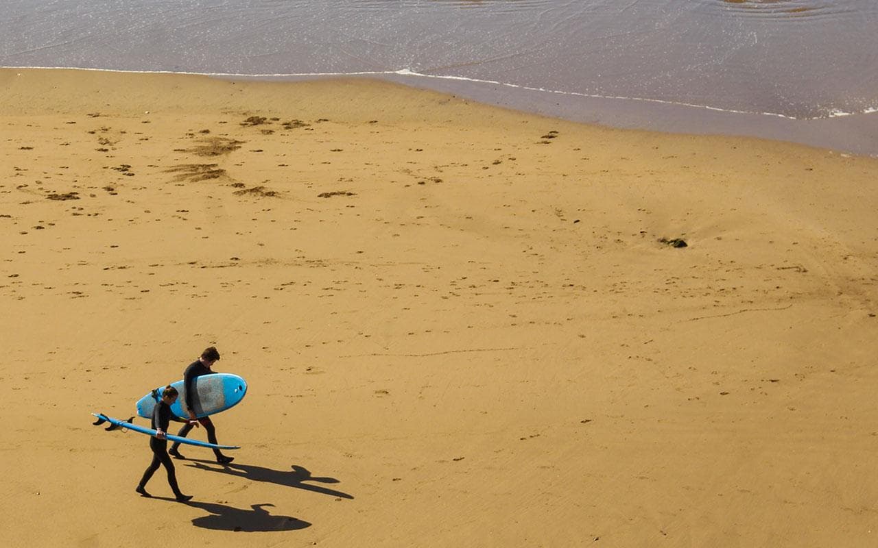 Two surfers carrying soft-top boards across the sand at Imsouane bay